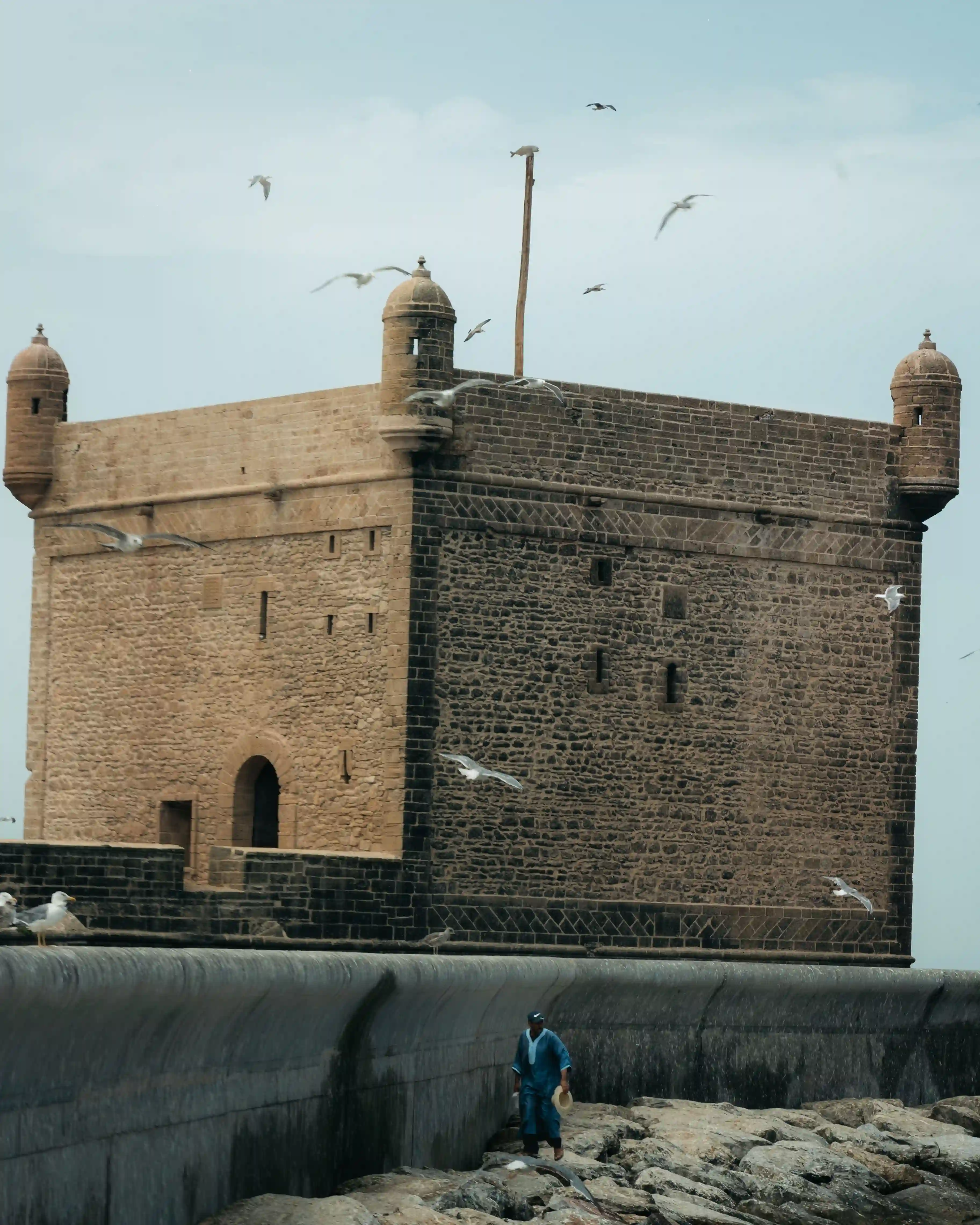 Tour en pierre de la Sqala du Port à Essaouira, entourée de mouettes en vol, avec une personne vêtue d'une djellaba bleue marchant le long de la digue au premier plan.
